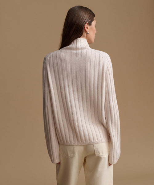 A woman shown from the back wearing a cream ribbed knit mock-neck sweater and matching cream trousers, set against a solid warm beige studio background.