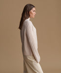 A woman in side profile wearing a cream ribbed knit mock-neck sweater and matching cream trousers, set against a solid warm beige studio background.