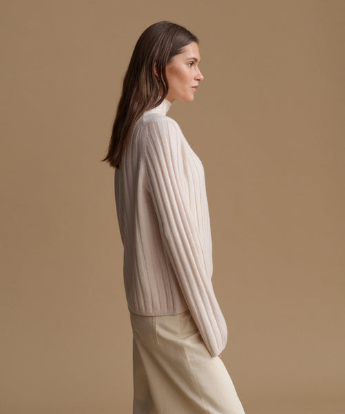 A woman in side profile wearing a cream ribbed knit mock-neck sweater and matching cream trousers, set against a solid warm beige studio background.