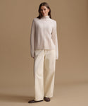 A woman stands against a warm beige studio background wearing a cream ribbed mock-neck sweater and matching cream wide-leg trousers, with dark brown shoes.