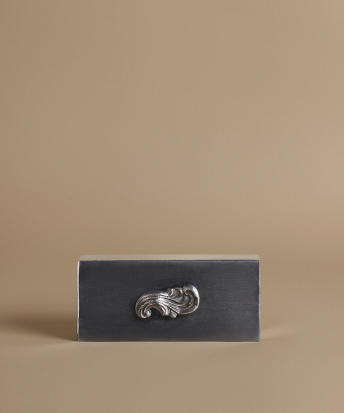 A rectangular dark charcoal gray box with a polished silver metallic scrollwork handle, set against a solid warm beige background.
