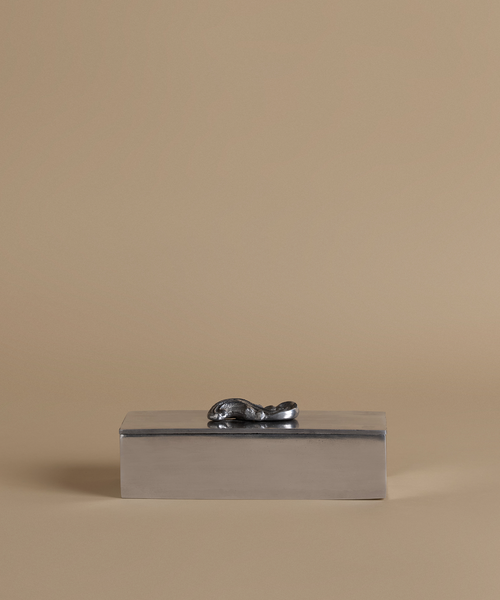 A rectangular metallic silver box with a small sculpted animal figure on top, set against a solid beige background.