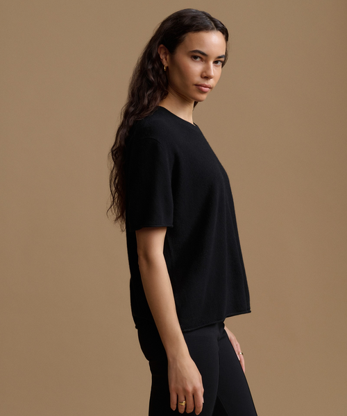 A woman  is shown in side profile wearing a black short-sleeve knit top and black pants against a solid warm tan studio background.