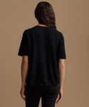 A woman viewed from the back wearing a plain black short-sleeve top and black pants against a warm tan studio background.