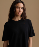 A woman wearing a solid black short-sleeve crewneck t-shirt, photographed against a solid warm beige studio background.