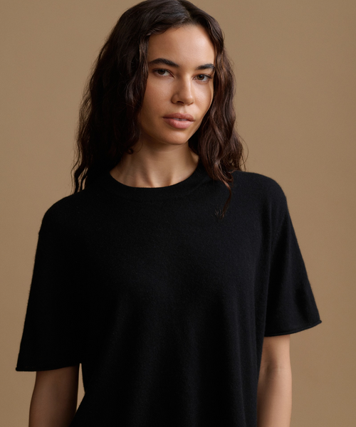 A woman wearing a solid black short-sleeve crewneck t-shirt, photographed against a solid warm beige studio background.