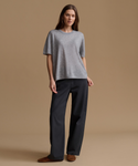 A woman stands against a plain beige studio background wearing a loose heather gray short-sleeve top, dark charcoal wide-leg pants, and brown slip-on shoes.