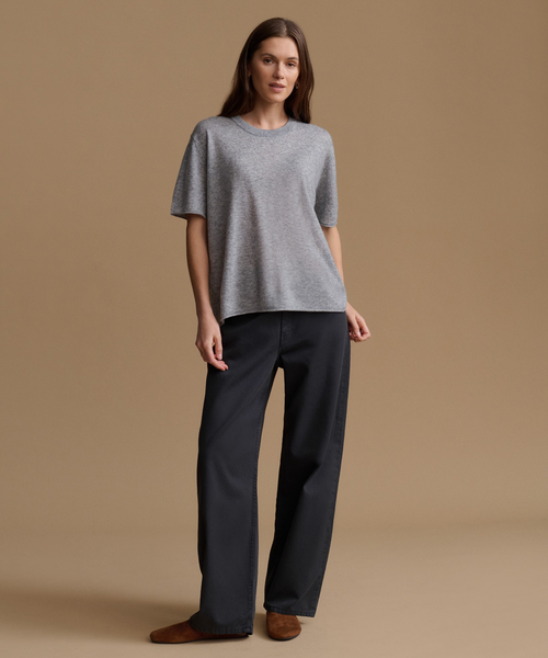 A woman stands against a plain beige studio background wearing a loose heather gray short-sleeve top, dark charcoal wide-leg pants, and brown slip-on shoes.