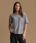 A woman modeling a short-sleeve crewneck t-shirt in heather gray, paired with black pants, against a solid tan studio background.