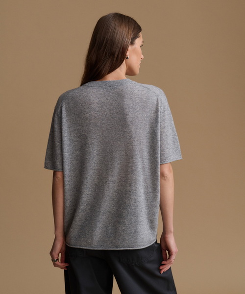 A woman shown from the back wearing a heather gray short-sleeve knit top and dark pants against a solid tan studio background.