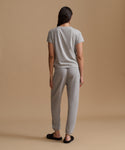 A woman viewed from behind wearing a light gray short-sleeve t-shirt and matching light gray jogger pants, standing against a solid beige studio background.