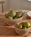 Two Seagrass Produce Bowls sit on a wooden table—one with artichokes, the other with pears. Plates, cutlery, and a glass nearby in soft natural light make these bowls perfect for stylish kitchen storage.