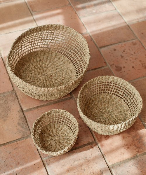 Three round Seagrass Produce Bowls in different sizes sit on a terracotta tiled floor. Made from natural fibers, theyre ideal as produce bowls or for stylish kitchen storage, featuring open, airy side weaving.