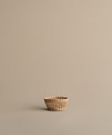 The Seagrass Produce Bowl rests empty on a plain, light beige surface with a matching background. Its minimal design and natural look make this bowl perfect for stylish, simple kitchen storage.
