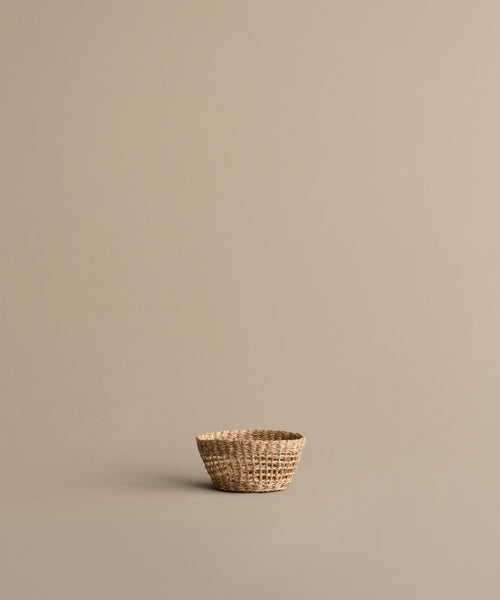 The Seagrass Produce Bowl rests empty on a plain, light beige surface with a matching background. Its minimal design and natural look make this bowl perfect for stylish, simple kitchen storage.