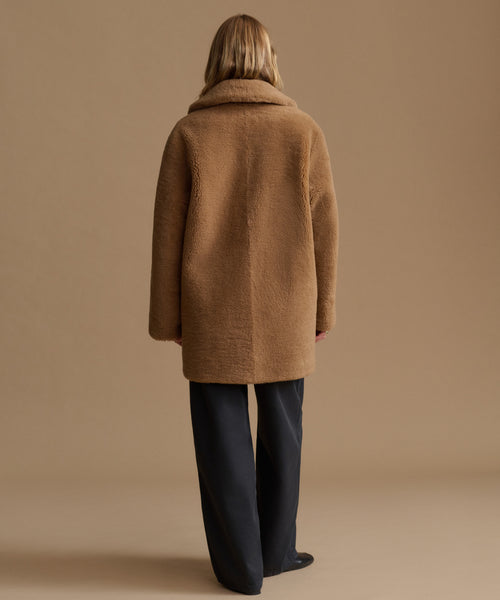 A woman viewed from behind wearing a camel-colored textured teddy coat and dark wide-leg trousers, standing against a solid warm beige studio background.