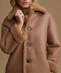 A close-up of a woman wearing a camel brown shearling-lined coat with three dark buttons, set against a solid warm beige background.