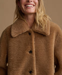 A close-up of a smiling woman wearing a warm camel brown sherpa fleece button-up jacket with a wide collar, set against a solid beige background.