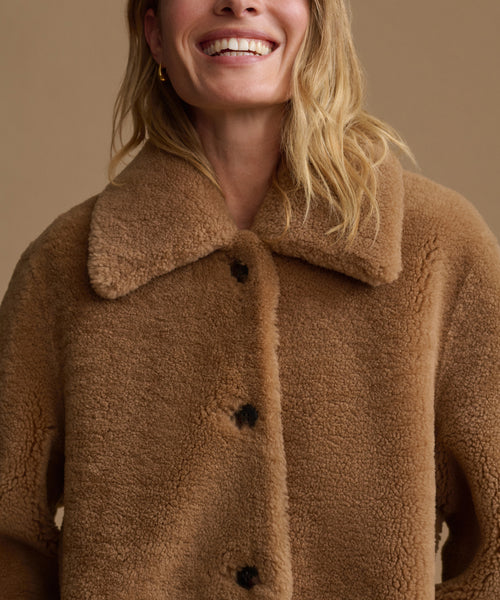 A close-up of a smiling woman wearing a warm camel brown sherpa fleece button-up jacket with a wide collar, set against a solid beige background.