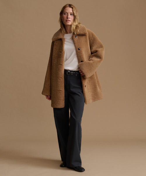 A woman stands against a warm beige studio background wearing a camel-colored teddy coat over a white top and dark charcoal trousers.