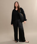 A woman stands against a beige background wearing the Simone V-Neck sweater in fine cotton merino, paired with wide-leg black satin pants, beige sandals, and a woven black tote—an effortless everyday look.