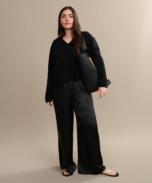 A woman stands against a beige background wearing the Simone V-Neck sweater in fine cotton merino, paired with wide-leg black satin pants, beige sandals, and a woven black tote—an effortless everyday look.