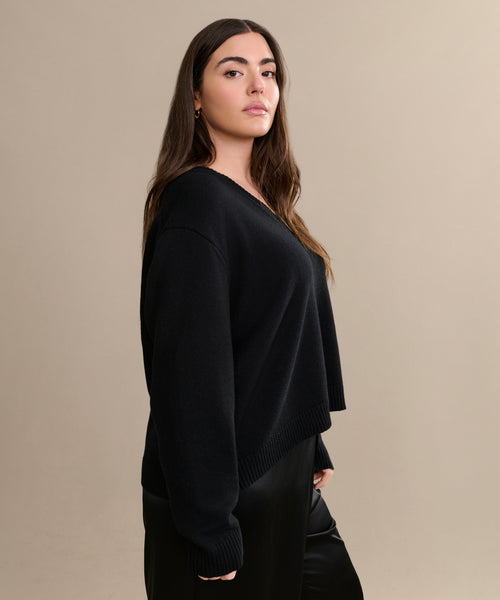 A woman with long brown hair wears the Simone V-Neck sweater in black fine cotton merino and black pants, standing confidently against a beige background—an effortless everyday layer.