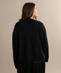 A woman with long brown hair is shown from behind, wearing the Simone V-Neck in black paired with black pants—an easy, everyday layer in fine cotton merino—standing against a plain beige background.