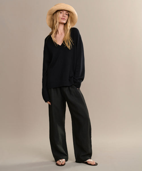 A woman stands relaxed against a neutral background, dressed in the Simone V-Neck—an everyday black fine cotton merino sweater—paired with wide-leg black pants, a straw hat, and sandals.