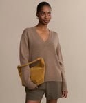 A woman wears the Simone V-Neck sweater in tan with olive green shorts and holds a mustard yellow suede clutch, standing against a plain beige background.