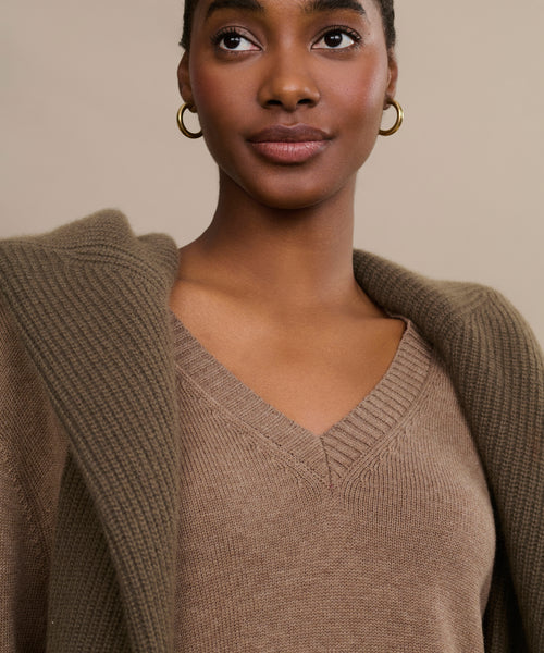 A person wears the Simone V-Neck sweater in brown cotton merino with a textured brown jacket over their shoulders and gold hoop earrings, set against a neutral background.