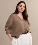 A woman with long brown hair exudes effortless style in the Simone V-Neck, a loose cotton-merino sweater, paired with white trousers and a brown belt, standing confidently against a beige background.