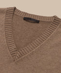 A close-up of the Simone V-Neck sweater, crafted from soft cotton merino with ribbed collar and neckline, is laid flat on a beige background. A black label is visible inside the collar.