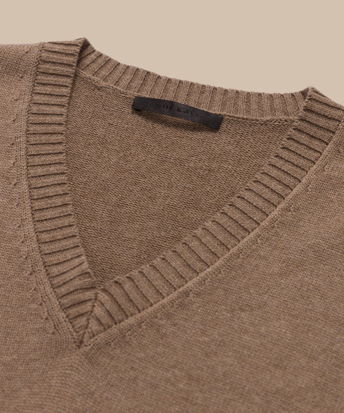 A close-up of the Simone V-Neck sweater, crafted from soft cotton merino with ribbed collar and neckline, is laid flat on a beige background. A black label is visible inside the collar.
