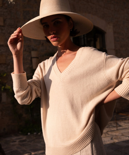 Wearing the Simone V-Neck, a person with a wide-brimmed hat stands outdoors in warm sunlight, their face shaded. Behind them, a stone building completes the scene.