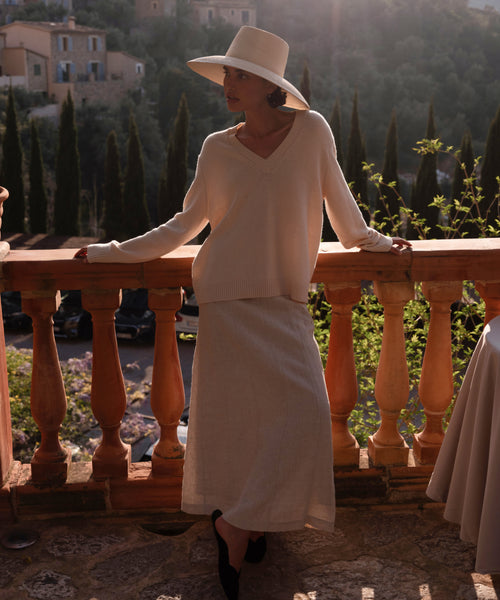 A woman in the Simone V-Neck ivory fine cotton merino sweater and long skirt stands by a stone railing, sunlight behind her, with trees and hills beyond—a perfect luxury layer for any season.