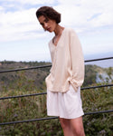A person with short brown hair wearing the Simone V-Neck in ivory fine cotton merino and white shorts stands outdoors by a railing, surrounded by greenery and a cloudy sky—a versatile luxury layer for any season.