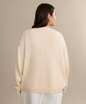 A woman with medium-length dark hair, silver hoop earrings, and a V-Neck Ivory Simone sweater with white pants stands facing away against a beige background—a perfect luxury layer.