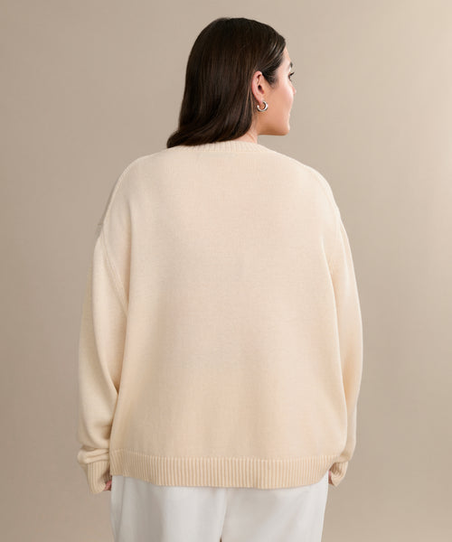 A woman with medium-length dark hair, silver hoop earrings, and a V-Neck Ivory Simone sweater with white pants stands facing away against a beige background—a perfect luxury layer.