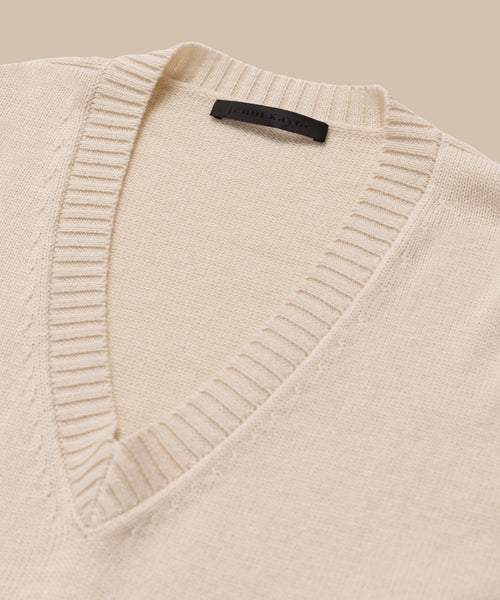 Simone V-Neck sweater in ivory, crafted from fine cotton-merino blend with a ribbed collar and black neck label, offers a touch of luxury—shown here on a beige background.