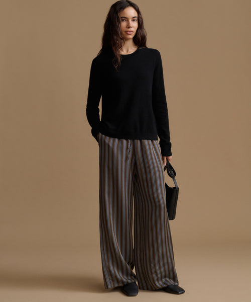 A model wearing high-waisted, wide-leg trousers with vertical brown, beige, and gray stripes, paired with a black long-sleeve top and black pointed shoes, set against a solid tan studio background.