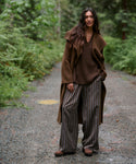 A woman stands on a gravel path in a forest wearing a brown long coat, brown knit sweater, brown-and-gray striped wide-leg pants, and brown shoes.