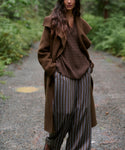 A woman stands on a gravel path in a forest wearing a brown long coat, brown knit sweater, brown-and-gray striped wide-leg pants, and brown shoes.
