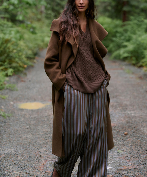 A woman stands on a gravel path in a forest wearing a brown long coat, brown knit sweater, brown-and-gray striped wide-leg pants, and brown shoes.