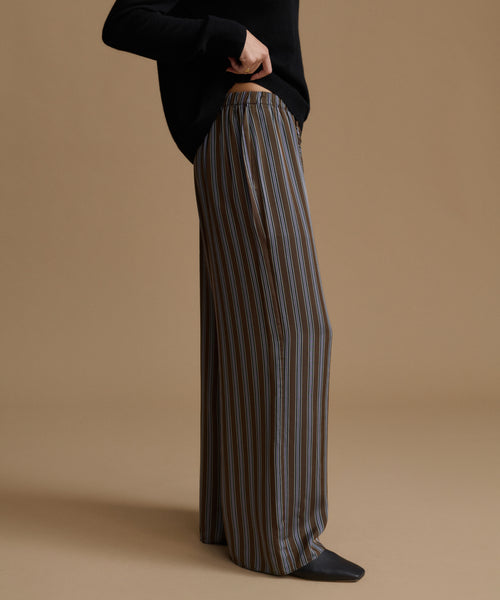 A model wearing high-waisted, wide-leg trousers with vertical brown, beige, and gray stripes, paired with a black long-sleeve top and black pointed shoes, set against a solid tan studio background.