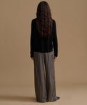A model wearing high-waisted, wide-leg trousers with vertical brown, beige, and gray stripes, paired with a black long-sleeve top and black pointed shoes, set against a solid tan studio background.
