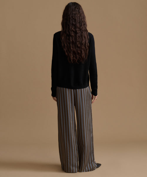 A model wearing high-waisted, wide-leg trousers with vertical brown, beige, and gray stripes, paired with a black long-sleeve top and black pointed shoes, set against a solid tan studio background.