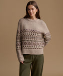A woman wearing a beige knit sweater with a brown and purple Fair Isle pattern across the chest and sleeves, paired with olive green pants, set against a solid beige background.