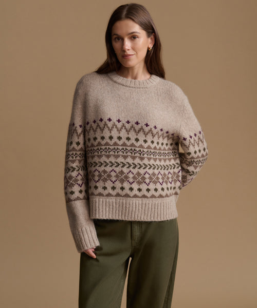 A woman wearing a beige knit sweater with a brown and purple Fair Isle pattern across the chest and sleeves, paired with olive green pants, set against a solid beige background.