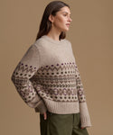 A woman wearing a beige knit sweater with a brown and purple Fair Isle pattern, paired with olive green pants, set against a solid tan studio background.